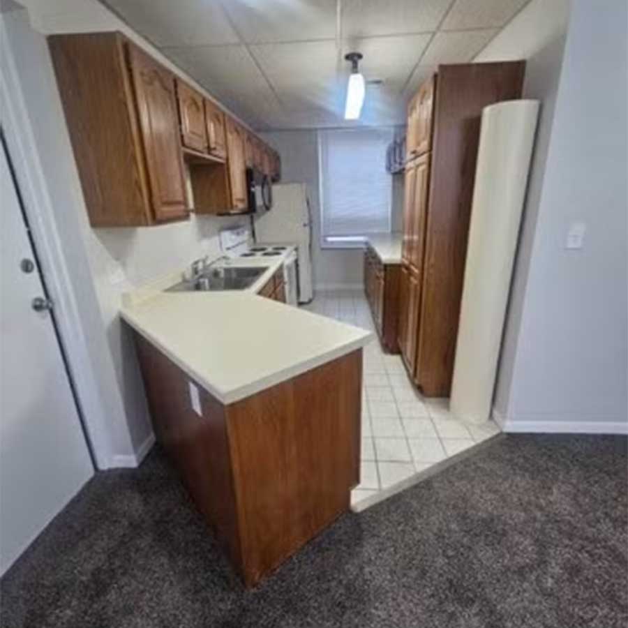 Small kitchen at Hereford & Hops Delta Apartments with wood cabinets and a carpeted area in front.
