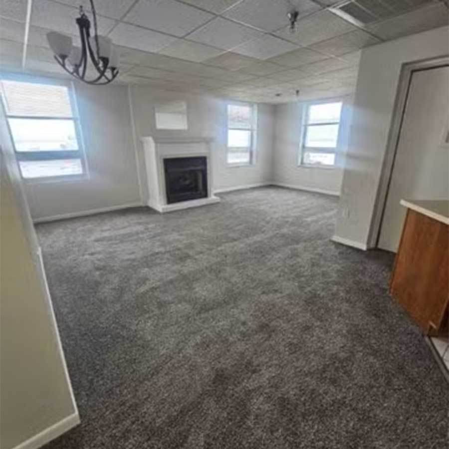 Empty carpeted living room at Hereford & Hops Delta Apartments with fireplace and partial kitchen counter.